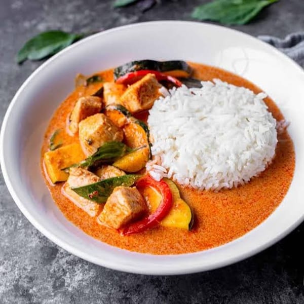 Red curry with chicken, zucchini, bell peppers, and white rice in a white bowl.