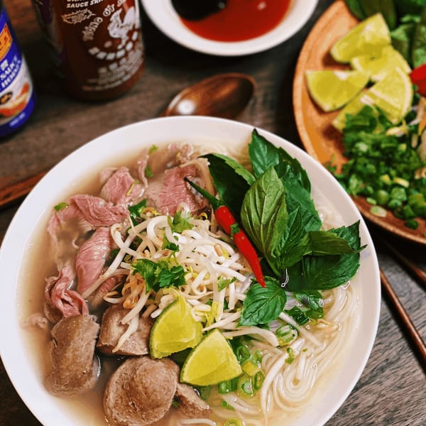 A bowl of pho with beef, meatballs, noodles, and fresh herbs, alongside Sriracha and hoisin sauce.