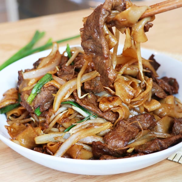 Beef chow fun with wide noodles, beef, onions, and green onions, lifted by chopsticks from a white bowl.